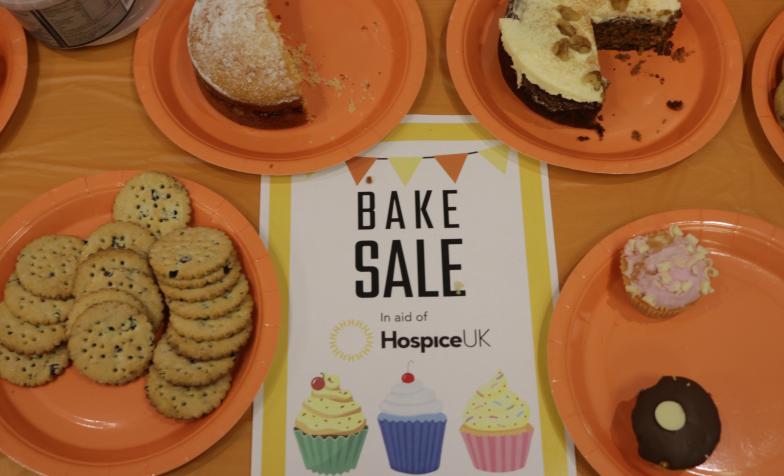 Bake Sale with an orange tablecloth and image of biscuits and cakes on plates surrounding a Hospice UK bake sale sign