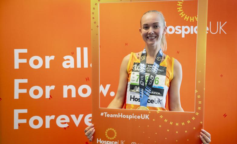Hospice UK runner smiling and holding a photo frame. She is stood in front of a sign which says 'For all. For now. Forever'.