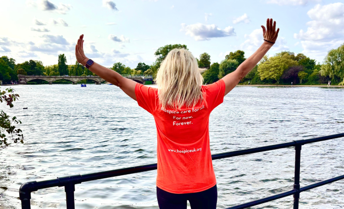 Woman wearing an orange Hospice UK tshirt with the words, 'Hospice Care for all. For Now. Forever.' She is stood in front of the Serpentine Lake with her arms spread wide.