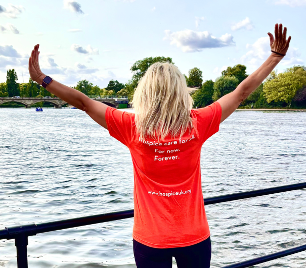 Woman wearing an orange Hospice UK tshirt with the words, 'Hospice Care for all. For Now. Forever.' She is stood in front of the Serpentine Lake with her arms spread wide.