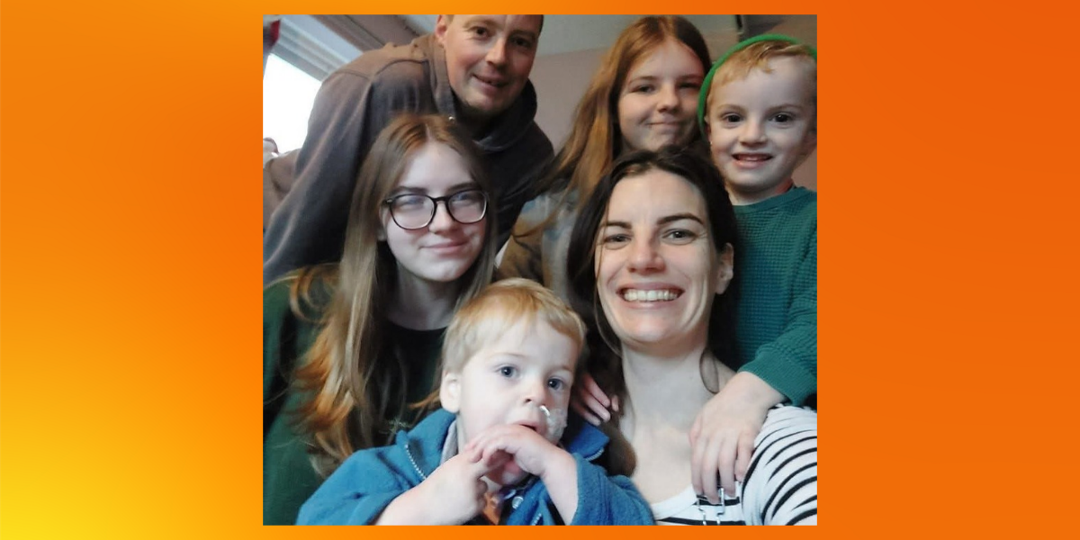 Caleb's family squeeze together for a smiling selfie. Caleb, in a blue jacket, sits at the front on his mum Jenna's lap, with his dad Jamie and three siblings leaning in around them.