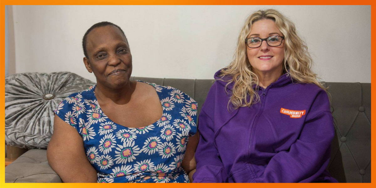 Dorcas and her care worker, Tracey, sit side by side on a grey sofa, smiling. Dorcas wears a blue dress with a pink and white flower pattern, and Tracey wears a purple Community Hospice hoodie.