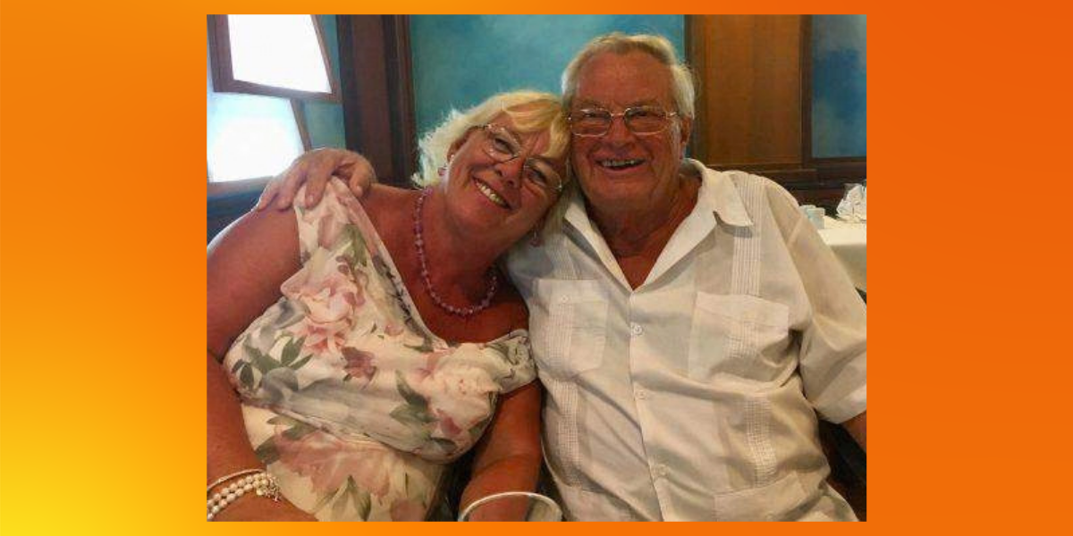 Michael and his wife, Rosemary, sit close together at a table, smiling, with his arm around her shoulders. Rosemary wears a floral dress and a pink beaded necklace, and Michael wears a white short-sleeved shirt and glasses.