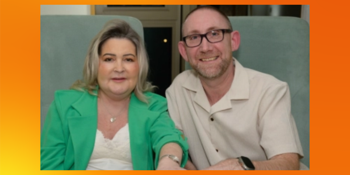 Michele and her husband, Frank, sit side by side in pale green armchairs, smiling. Michele wears a green blazer over a white top, and Frank wears a cream short-sleeved shirt and glasses.