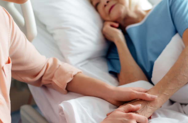 A patient in a hospital bed, with someone sitting by their bedside holding their hand