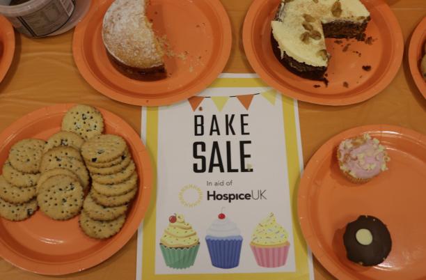 Bake Sale with an orange tablecloth and image of biscuits and cakes on plates surrounding a Hospice UK bake sale sign