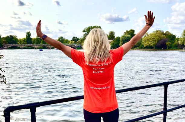 Woman wearing an orange Hospice UK tshirt with the words, 'Hospice Care for all. For Now. Forever.' She is stood in front of the Serpentine Lake with her arms spread wide.
