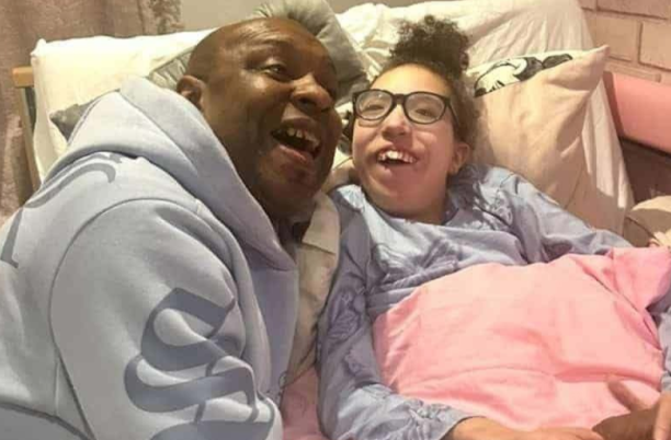 A dad is smiling next to his daughter, who is lying in bed. They both look very happy.