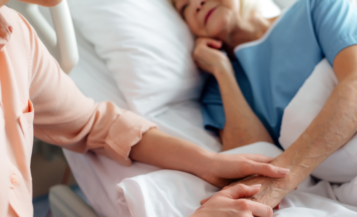 A patient in a hospital bed, with someone sitting by their bedside holding their hand