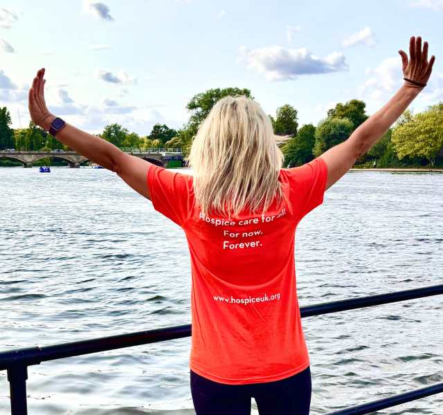 Woman wearing an orange Hospice UK tshirt with the words, 'Hospice Care for all. For Now. Forever.' She is stood in front of the Serpentine Lake with her arms spread wide.