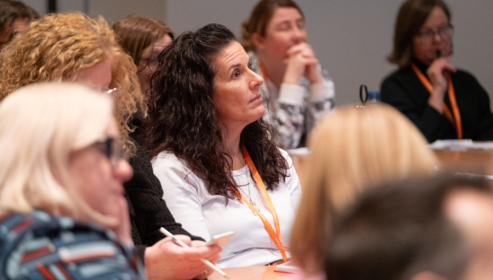 A woman with long dark hair sitting in a crowded room at a conference event.