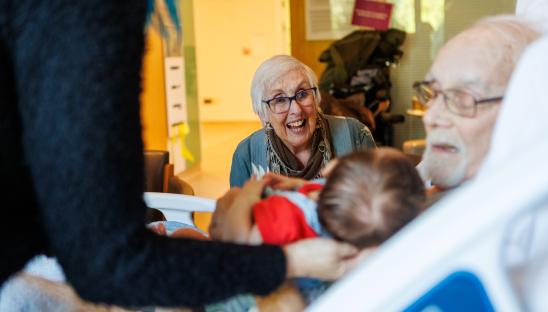 Family gathered around man in a bed. A woman is smiling at the baby he is holding.